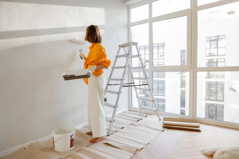 Woman painting interior wall in a bright, modern apartment with large windows.